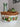 A round white ceramic bowl with a brown woven wicker handle and rim, decorated with hand-painted brown and green floral motifs, displayed on a white surface with decorative glass vases in the background.