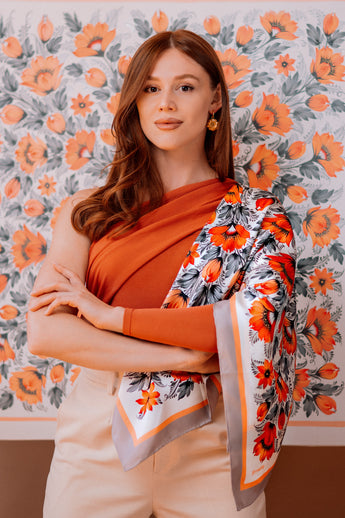 Woman wearing an orange top and floral scarf against a floral patterned wall.