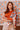 Woman wearing an orange top and floral scarf against a floral patterned wall.